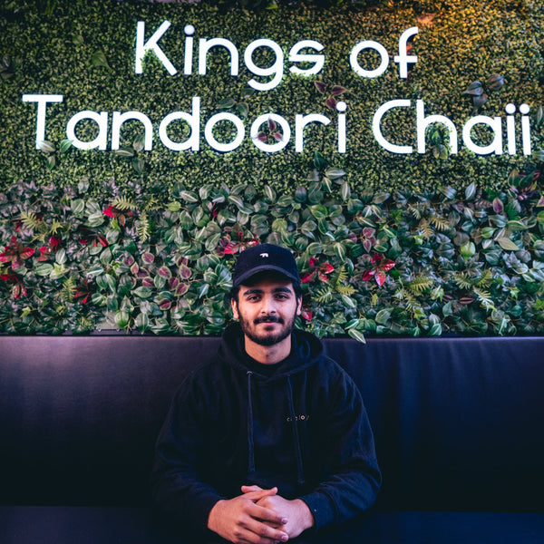 Person sitting on a couch in front of a wall with 'Kings of Tandoori Chaii' text and greenery.